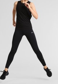 Černý sportovní top bez rukávů a těsné legíny. Hladký materiál, bez rukávů. Černé tenisky s tkaničkami. Dynamická póza s jednou nohou nataženou.