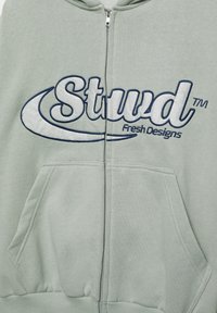Felpa con cappuccio verde chiaro con zip, tasca frontale, polsini elastici e logo ricamato "Stud" in blu scuro. Tessuto morbido.