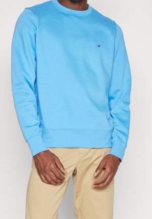 Man wearing light blue long-sleeve sweatshirt and beige pants, hands relaxed near hips, against plain light background.