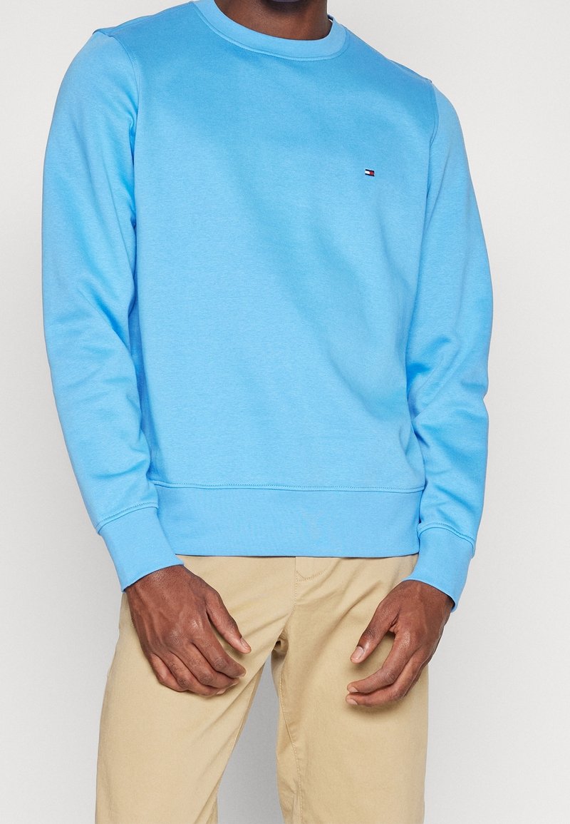 Homme portant un sweat-shirt à manches longues bleu clair et un pantalon beige, les mains détendues près des hanches, sur un fond clair uni.