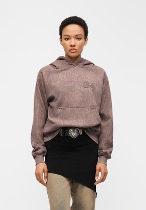 Femme portant un sweat à capuche mauve avec un texte brodé, une superposition de jupe asymétrique noire, un pantalon beige et une ceinture avec une boucle en forme de cœur.