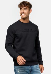 Sudadera negra con cuello redondo y lettering texturizado "INDICODE JEANS" en la parte delantera. Presenta puños y dobladillo acanalados.