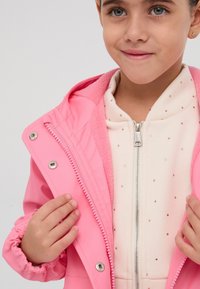 Giacca impermeabile rosa con zip frontale, hardware argentato e polsini elasticizzati, indossata sopra una maglietta di colore chiaro con piccole decorazioni.