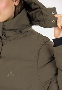 Braune isolierte Jacke mit hohem Kragen, Druckknöpfen und einem Logo. Verfügt über eine glatte Textur und ein strukturiertes Design für Wärme.