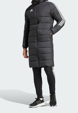 Homme portant une longue veste matelassée noire avec capuche, des rayures blanches Adidas sur les manches, un pantalon de survêtement noir Adidas, et des baskets noires avec semelles blanches.