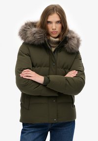 Chaqueta acolchada de color verde oliva con capucha forrada de pelo, textura acolchada, bolsillos frontales y cierre de botones, llevada sobre un suéter beige.