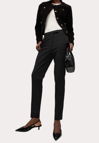 Czarna teksturowana krótka kurtka z złotymi guzikami, zestawiona z dopasowanymi czarnymi spodniami i czarnymi szpilkami na odkrytej pięcie, trzymając czarną torebkę.