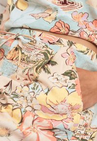 Primer plano de una persona que lleva ropa con estampado colorido de flores y paisajes, con una mano apoyada en un bolsillo lateral.