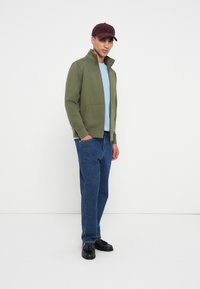 Χακί ζακέτα με φερμουάρ και ψηλό γιακά, ανοιχτό μπλε μπλούζα, μπλε τζιν και μαύρα παπούτσια. Μπουργκουντί καπέλο προσθέτει χρωματική έμφαση; το μοντέλο στέκεται προφίλ.