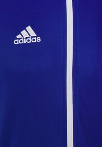 Blå zip-jacka i slät tyg med vit dragkedja och Adidas-logotyp i vitt på vänster bröst.