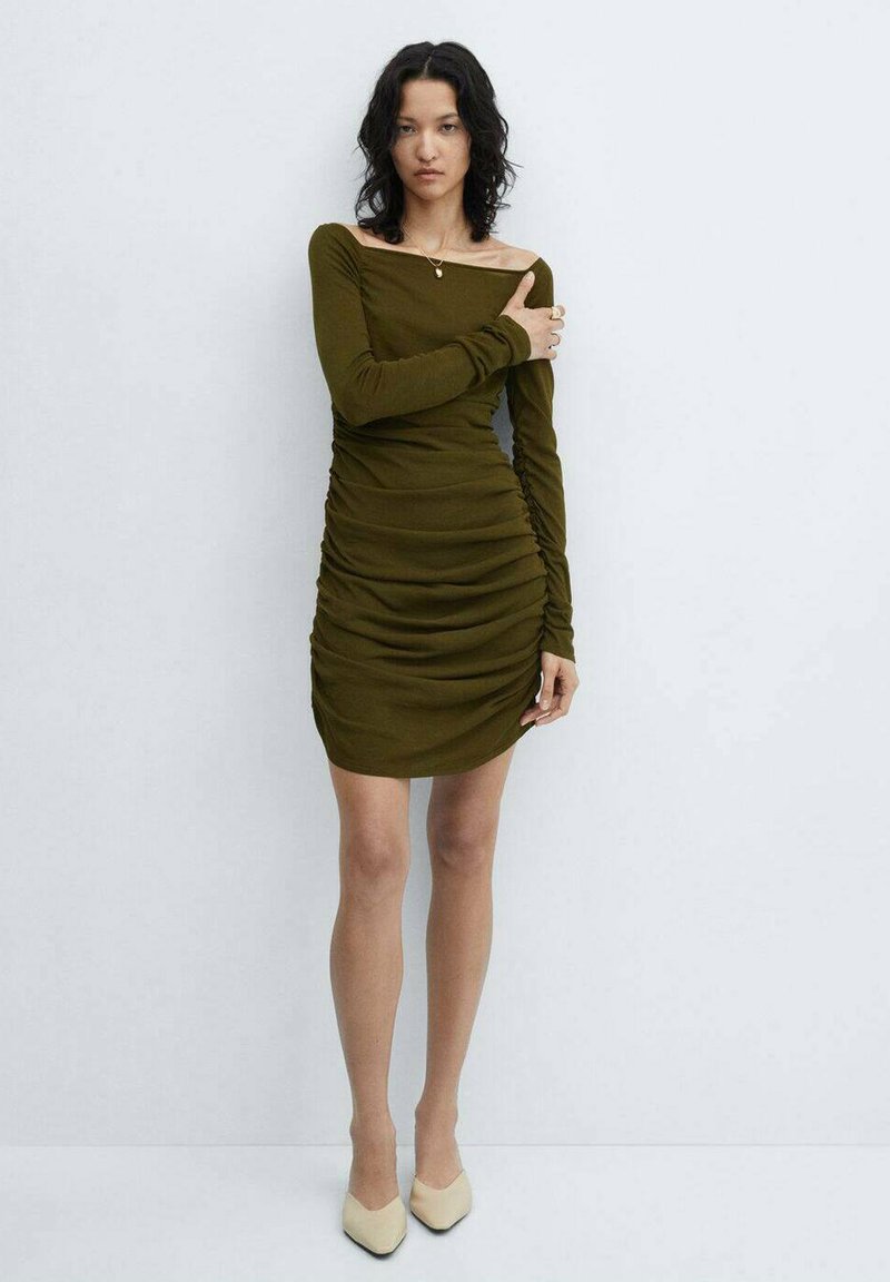 Olivno zelena obleka z dolgimi rokavi, z nabranimi detajli, prilegajočim krojem in izrezom izpod ramen. Obuta v svetle čevlje s koničastim nosom.