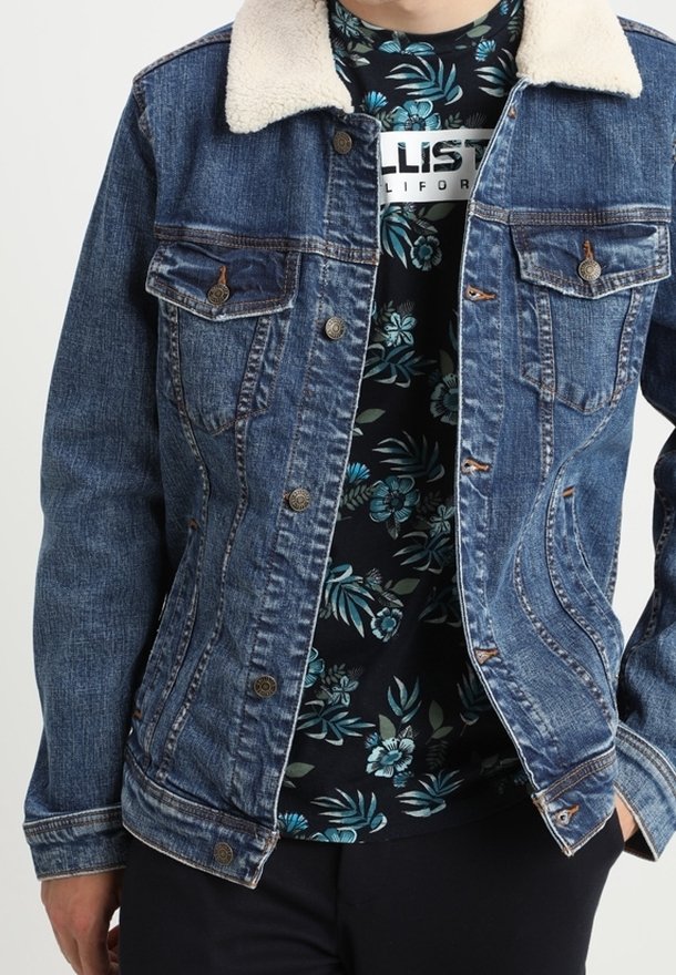 Jeansjacka med blåtvätt, fuskpälskrage och två bröstfickor. Underneath syns en svart skjorta med blommönster.