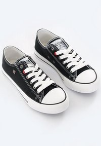 Une paire de baskets basses en toile noires avec des semelles en caoutchouc blanches, des renforts aux orteils, des lacets blancs et une étiquette de marque visible sur les languettes.