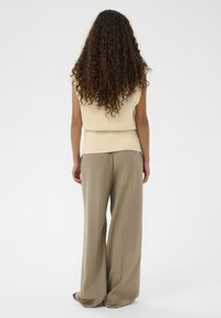 Frau mit langen lockigen Haaren, die ein ärmelloses cremefarbenes Oberteil und weite beigen Hosen trägt, stehend und mit dem Rücken zur Kamera vor einem schlichten Hintergrund.