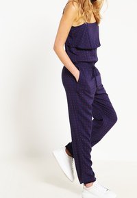 Marinblå jumpsuit med volangtop, röd blommönster, elastisk midja, sidofickor och tapered ben, kombinerad med vita sneakers.