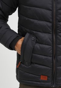 Main dans la poche boutonnée d'une veste matelassée noire avec fermeture éclair frontale et une petite étiquette de marque en cuir marron près de l'ourlet.