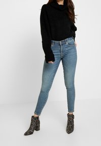 Černý svetr s texturovaným pletením a vysokým límcem, doplněný o světle modré, těsně padnoucí džíny a černo-bílé kotníkové boty se vzorem hada.