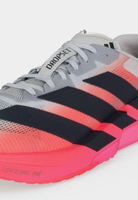 Γκρι και νεον ροζ αθλητικό παπούτσι Adidas με μαύρες ρίγες, λευκά κορδόνια, ετικέτα "DROPS" στη γλώσσα και "LIGHTSTRIKE PRO" στην άκρη της σόλας.