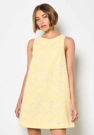 Młoda kobieta o krótkich włosach, ubrana w żółtą sukienkę bez rękawów z białymi kwiatowymi wzorami, stojąca na tle jednolitego białego tła.