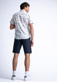 Camisa de manga corta en blanco con un patrón de dibujos a línea negros, combinada con shorts marinos. El modelo lleva zapatillas blancas con detalles verdes.