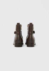 Brune læderankelstøvler med en glat finish, sidelommer med lynlås og en rem med spænde. Har lave stabelhæl. Set bagfra.