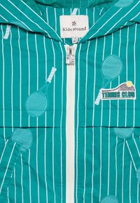 Παιδικό μπουφάν σε τιρκουάζ με λευκές κάθετες ρίγες, σχέδια με ρακέτες τένις, λευκό φερμουάρ και πατς με την επιγραφή "Tennis Club" στο μπροστινό μέρος.