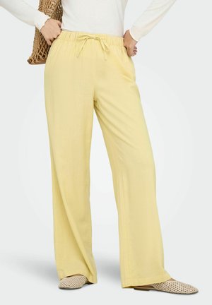 Kobieta nosząca luźne żółte spodnie ze ściągaczem, biały top z długim rękawem, beżowe plecione buty wsuwane i plecioną brązową torbę na ramię.