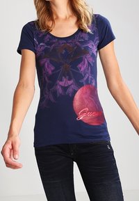 Mujer vistiendo una camiseta ajustada de manga corta azul marino con un diseño floral púrpura y un círculo rosa con texto en cursiva en la parte frontal, combinada con jeans oscuros.