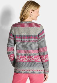 Eine Frau trägt einen mehrfarbigen Strickpullover mit geometrischen und floralen Mustern in Pink, Grau und Weiß, kombiniert mit rosa Hosen.