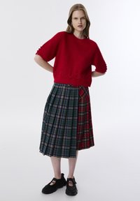 Roter Strickpullover mit kurzen, puffigen Ärmeln, der ein strukturiertes Logo zeigt. Darunter ein mehrfarbiges, gemustertes, plissiertes Kleid mit Karos in grünen und roten Tönen.