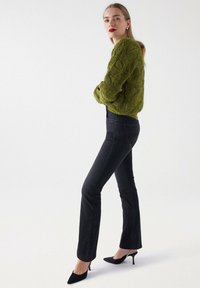 Suéter verde de punto con patrón texturizado, jeans negros acampanados y tacones negros de punta. La modelo se encuentra de perfil contra un fondo neutro.