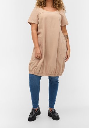 Beiges Baumwollkleid mit kurzen Ärmeln, rundem Ausschnitt, Seitentaschen und elastischem Saum. Über blauen engen Jeans und schwarzen Mokassins getragen.