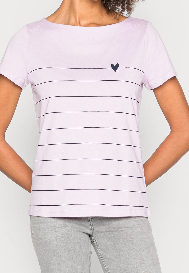 Femme portant un t-shirt rose clair à manches courtes avec de fines rayures noires horizontales et un petit cœur noir près de la poitrine, associé à un pantalon gris.