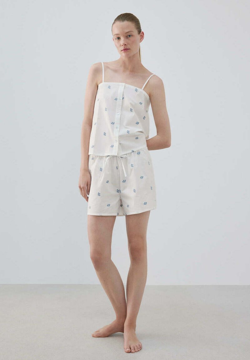 Jeune femme debout pieds nus portant un ensemble de pyjama blanc avec de fines bretelles et de petites impressions florales bleues sur le haut et le short.