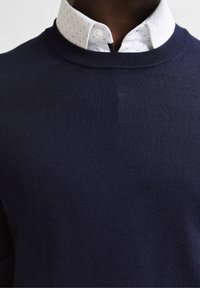 Marinblå stickad tröja med en slät textur, prydd med en vit kragskjorta under med ett subtilt mönster av små prickar.