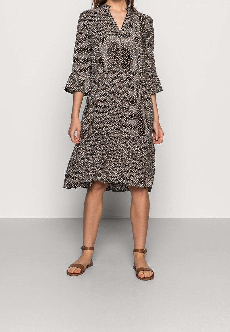 Černé a béžové šaty se vzorem, tříčtvrtečními rukávy, výstřihem do V a rozšířenou sukní. Doplněné hnědými sandály na pásky.