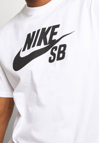 Vit t-shirt i bomull med en framträdande svart Nike-logotyp och "SB"-text, rund halsringning, korta ärmar och slät textur.