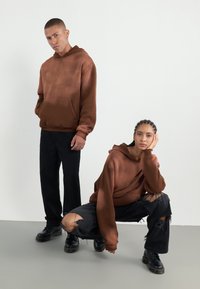 Hanorace cu glugă de culoare maro, cu un design gradient, purtate cu blugi negri rupți și bocanci negri, pe un fundal neutru.