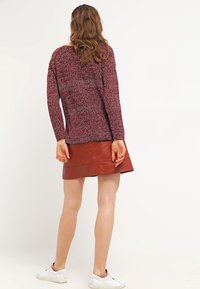 Pitkähiuksinen nainen pukeutuneena viininpunaiseen neulepuseroon, ruskeaan nahkahameeseen ja valkoisiin lenkkareihin, seisomassa yksiväristä valkoista taustaa vasten.