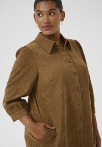 Woman with dark hair wearing hoop earrings and a brown corduroy button-up dress with three-quarter sleeves, looking to her left.