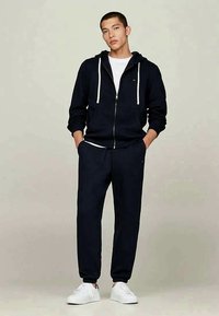 Marinblå zip-up hoodie med vita dragskor, matchande joggers. Buren över en vit t-shirt och vita sneakers. Slät tygstruktur.