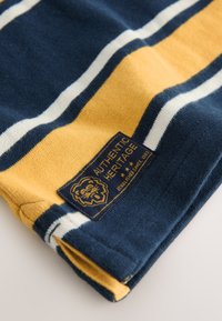 Tissu rayé en bleu marine, jaune et blanc. Présente une étiquette avec "Authentic Heritage" et un logo de lion. Texture douce en maille.