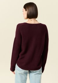 Burgundy tröja i mjukt stickat material med avslappnad passform, bateau-halsringning och långa ärmar. Kombinerad med ljusblå jeans.