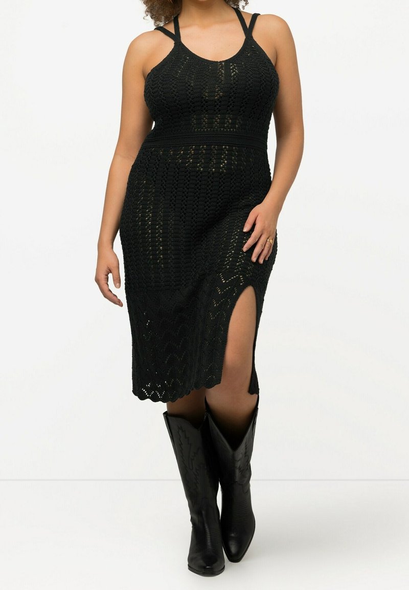 Femme portant une robe midi en crochet noire avec une fente à la cuisse et des bottes de cowboy noires pointues, sur un fond blanc uni.