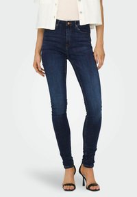 Žena nosi tamnoplave skinny traperice visokog struka u kombinaciji s crnim sandalama na visoku petu i bijelom kratkom jaknom preko bež majice.