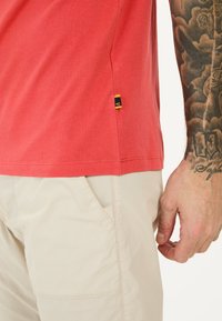 Rød bomulds-t-shirt med korte ærmer, med et side-hæm label. Beige shorts synlige, der viser en hånd med tatoveringer.