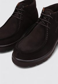 Botas de ante marrón hasta el tobillo con puntera redondeada, diseño de cordones, costuras en contraste y suela de goma gruesa y texturizada.