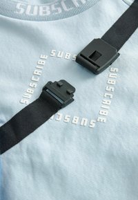 Gaiši zils audums ar uzrakstu "PARAKSTIES" baltā krāsā. Iezīmēti ar melnām siksnām un plastmasas piederumiem, lai varētu pielāgot izmēru.