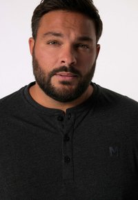 Tummanharmaa henley-paita, jossa on teksturoitu kangas, kolme nappia ja pieni logo rinnassa. Lähikuva paidasta.