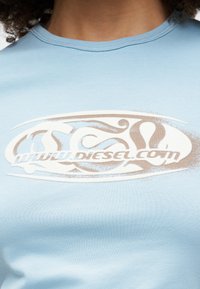 T-shirt azul claro feito de algodão suave, com um design de logotipo branco e castanho no peito em um padrão ondulado.